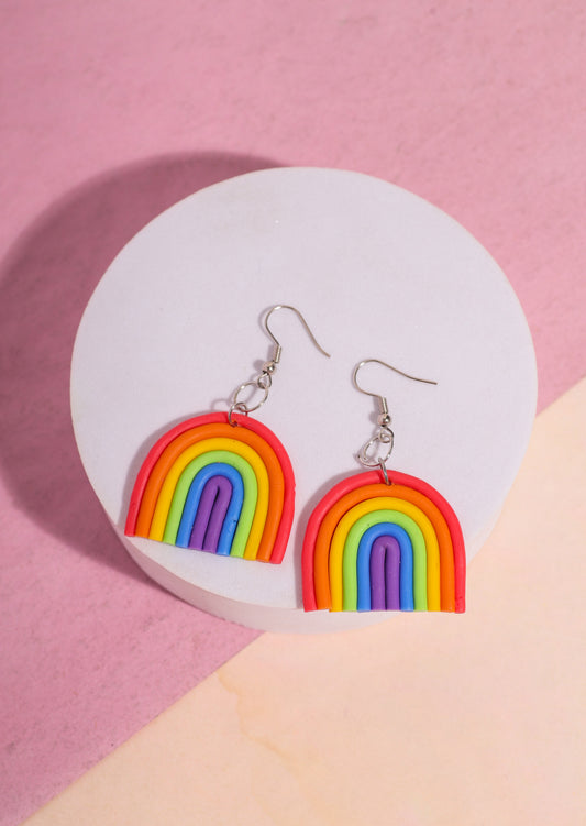 A pair of rainbow-shaped polymer clay earrings placed on a pink and beige background. The earrings feature layered arcs in red, orange, yellow, green, blue, and purple with silver hooks.
