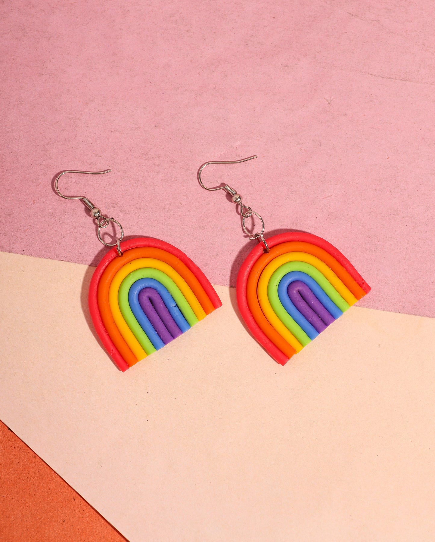 A pair of rainbow-shaped polymer clay earrings placed on a pink and beige background. The earrings feature layered arcs in red, orange, yellow, green, blue, and purple with silver hooks.