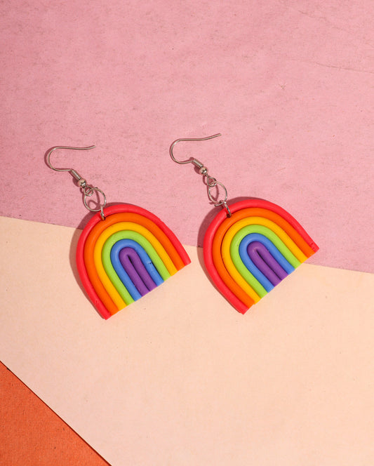 A pair of rainbow-shaped polymer clay earrings placed on a pink and beige background. The earrings feature layered arcs in red, orange, yellow, green, blue, and purple with silver hooks.