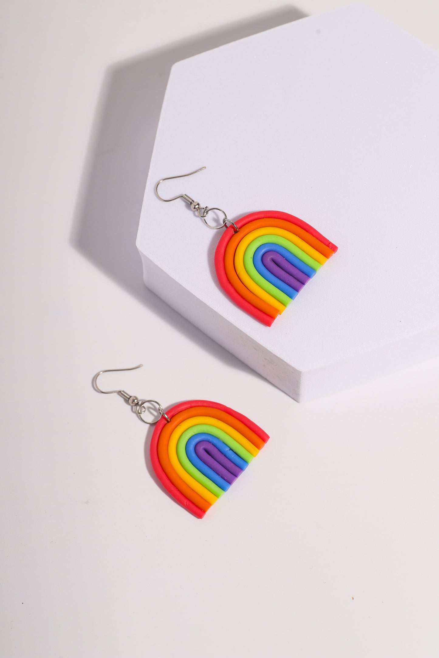 A pair of rainbow-shaped polymer clay earrings placed on a pink and beige background. The earrings feature layered arcs in red, orange, yellow, green, blue, and purple with silver hooks.