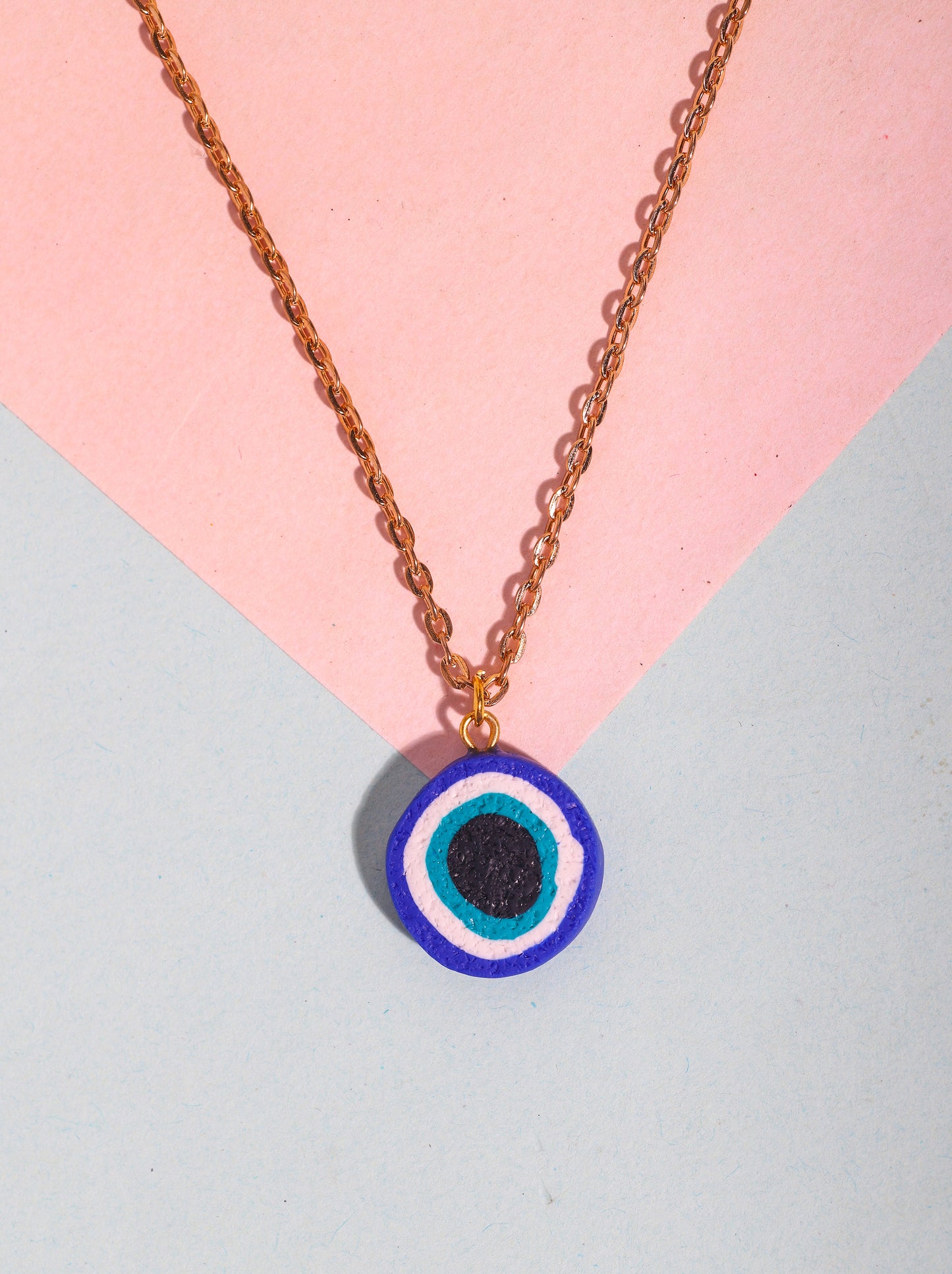 Close-up of a handmade evil eye necklace with a polymer clay charm on a gold-toned chain, styled on a soft pastel pink and blue background.