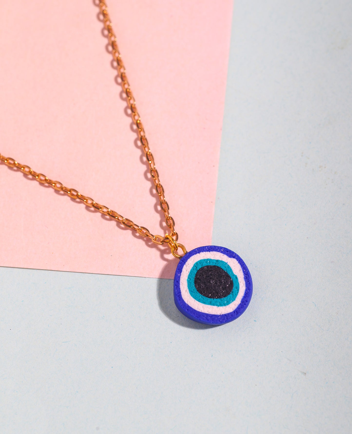 Close-up of a handmade evil eye necklace with a polymer clay charm on a gold-toned chain, styled on a soft pastel pink and blue background.