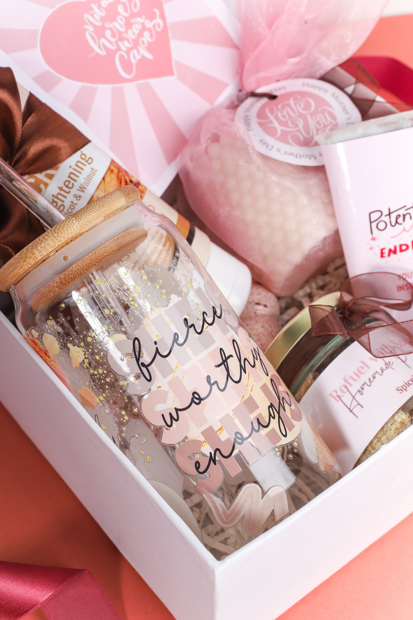 Empowerment-themed gift box featuring a pink chocolate bar with inspirational message, bamboo-lid glass tumbler with ‘fierce, worthy, enough’ quote, facial scrub tube, white bubble candle, jar of nuts, greeting card, and pink satin scrunchie on a peach background.