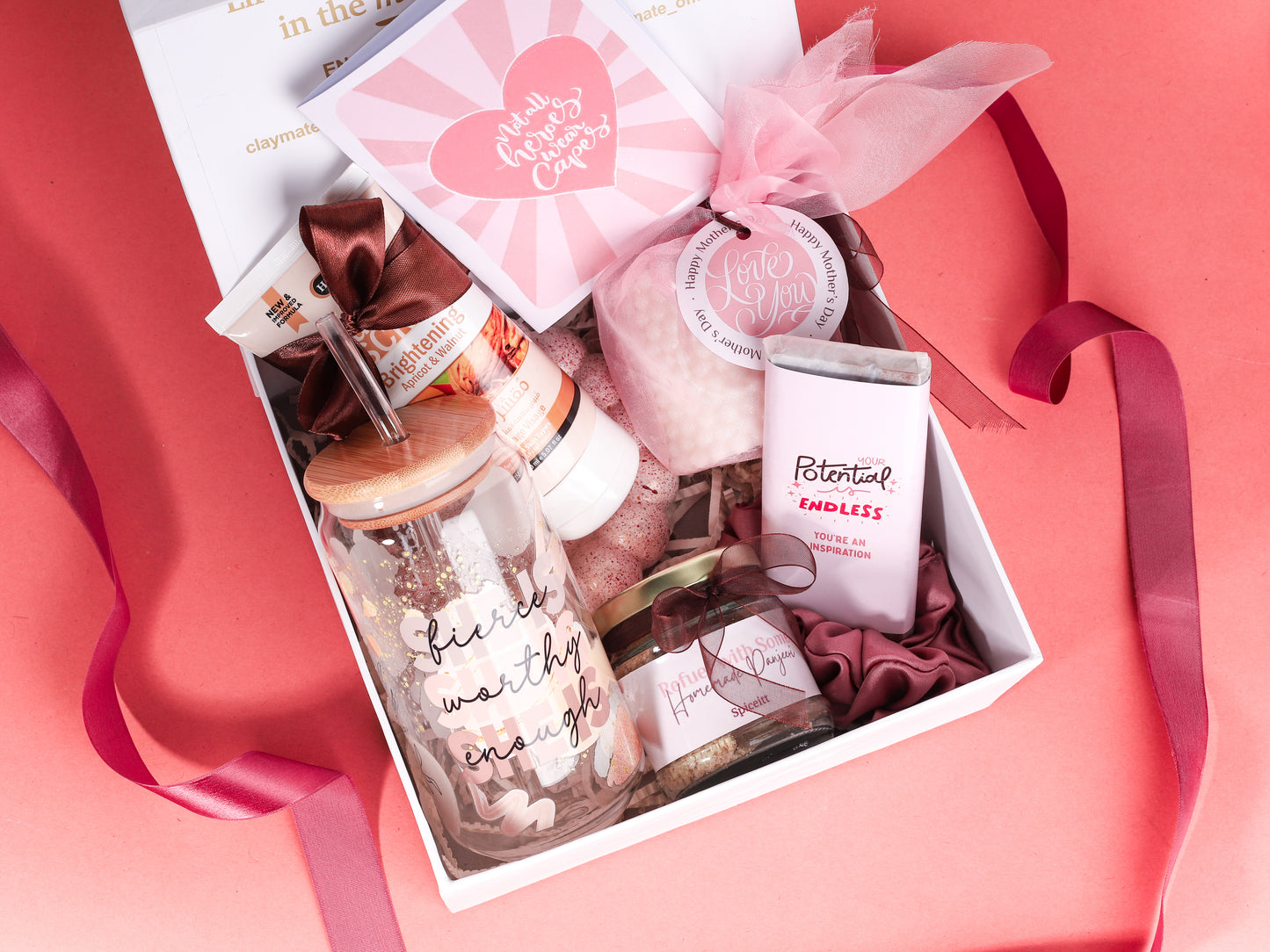 Empowerment-themed gift box featuring a pink chocolate bar with inspirational message, bamboo-lid glass tumbler with ‘fierce, worthy, enough’ quote, facial scrub tube, white bubble candle, jar of nuts, greeting card, and pink satin scrunchie on a peach background.