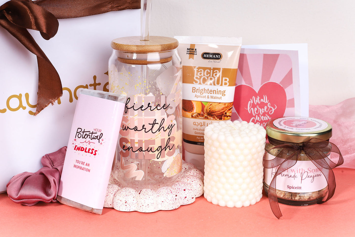 Empowerment-themed gift box featuring a pink chocolate bar with inspirational message, bamboo-lid glass tumbler with ‘fierce, worthy, enough’ quote, facial scrub tube, white bubble candle, jar of nuts, greeting card, and pink satin scrunchie on a peach background.