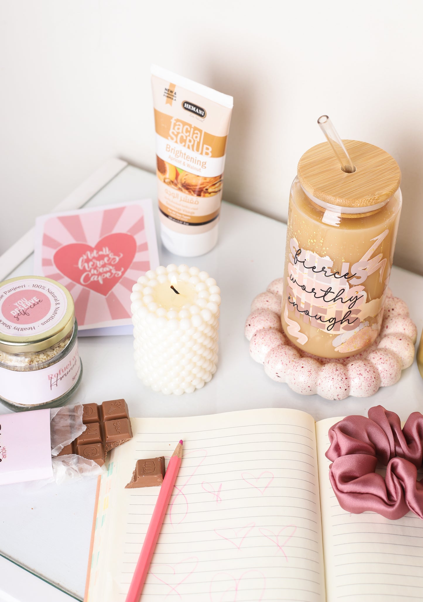 Empowerment-themed gift box featuring a pink chocolate bar with inspirational message, bamboo-lid glass tumbler with ‘fierce, worthy, enough’ quote, facial scrub tube, white bubble candle, jar of nuts, greeting card, and pink satin scrunchie on a peach background.