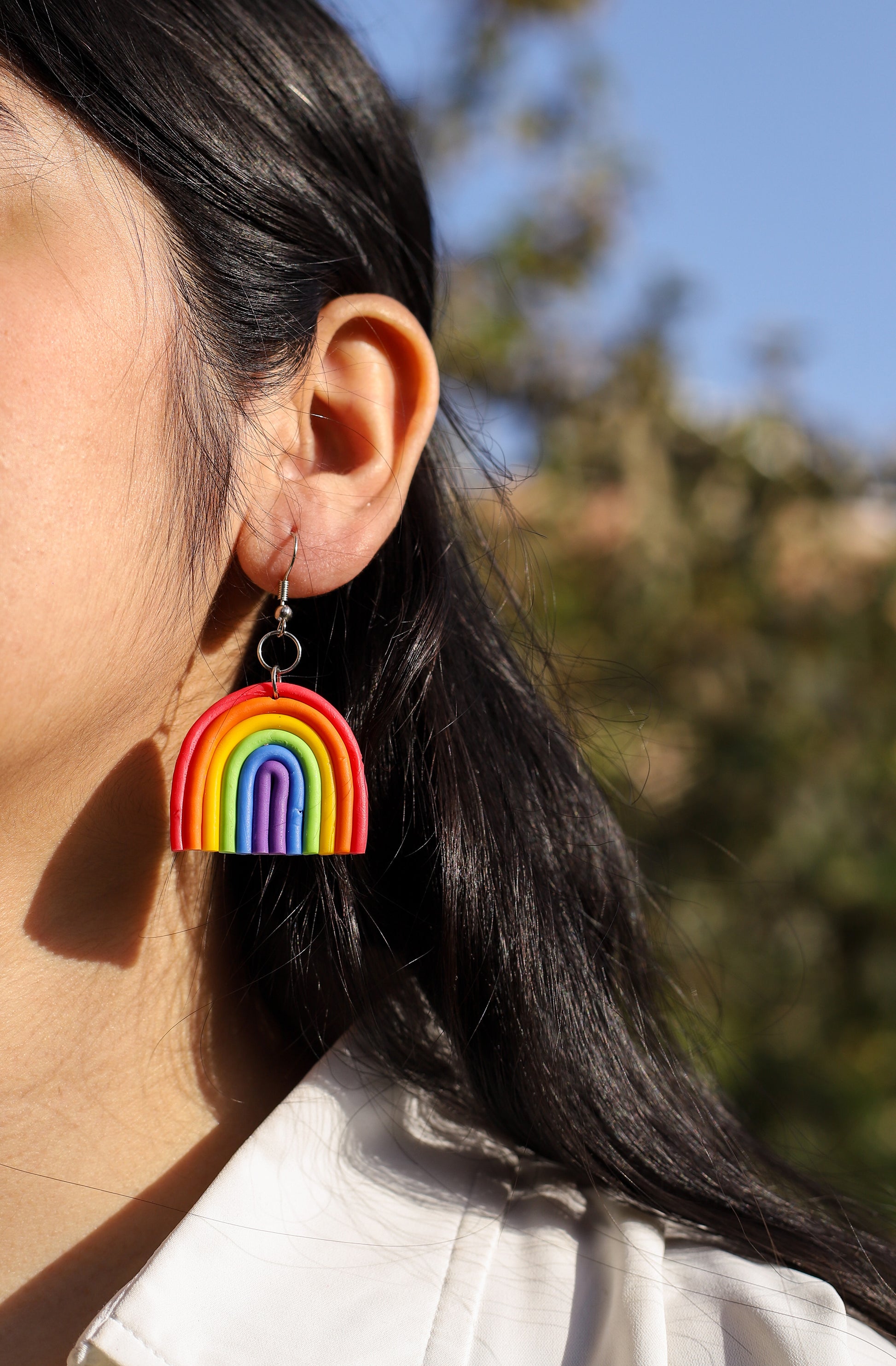 Girl wearing rainbow-shaped polymer clay earrings. The earrings feature layered arcs in red, orange, yellow, green, blue, and purple with silver hooks.