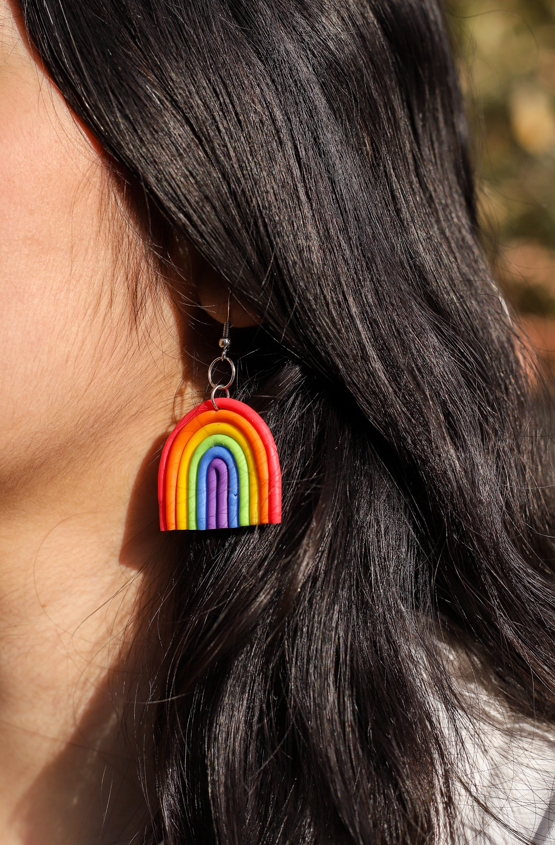 Girl wearing rainbow-shaped polymer clay earrings. The earrings feature layered arcs in red, orange, yellow, green, blue, and purple with silver hooks.
