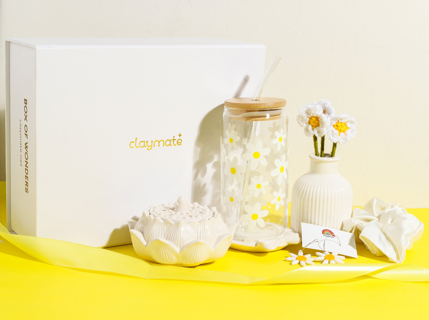 White gift box with yellow daisy-themed accessories including a glass tumbler with bamboo lid, crochet flower stems in a vase, ceramic trinket dish, daisy earrings, and a white scrunchie on a bright yellow background, Claymate gift set.