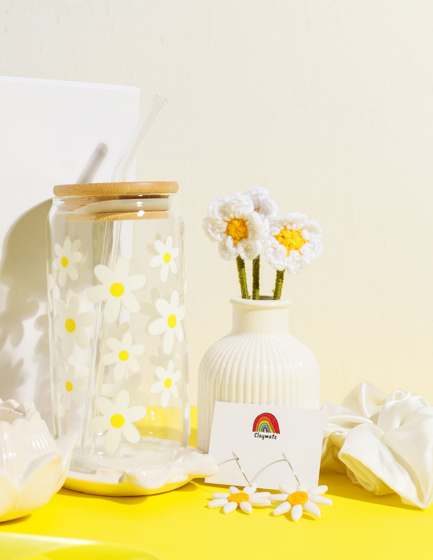 White gift box with yellow daisy-themed accessories including a glass tumbler with bamboo lid, crochet flower stems in a vase, ceramic trinket dish, daisy earrings, and a white scrunchie on a bright yellow background, Claymate gift set.