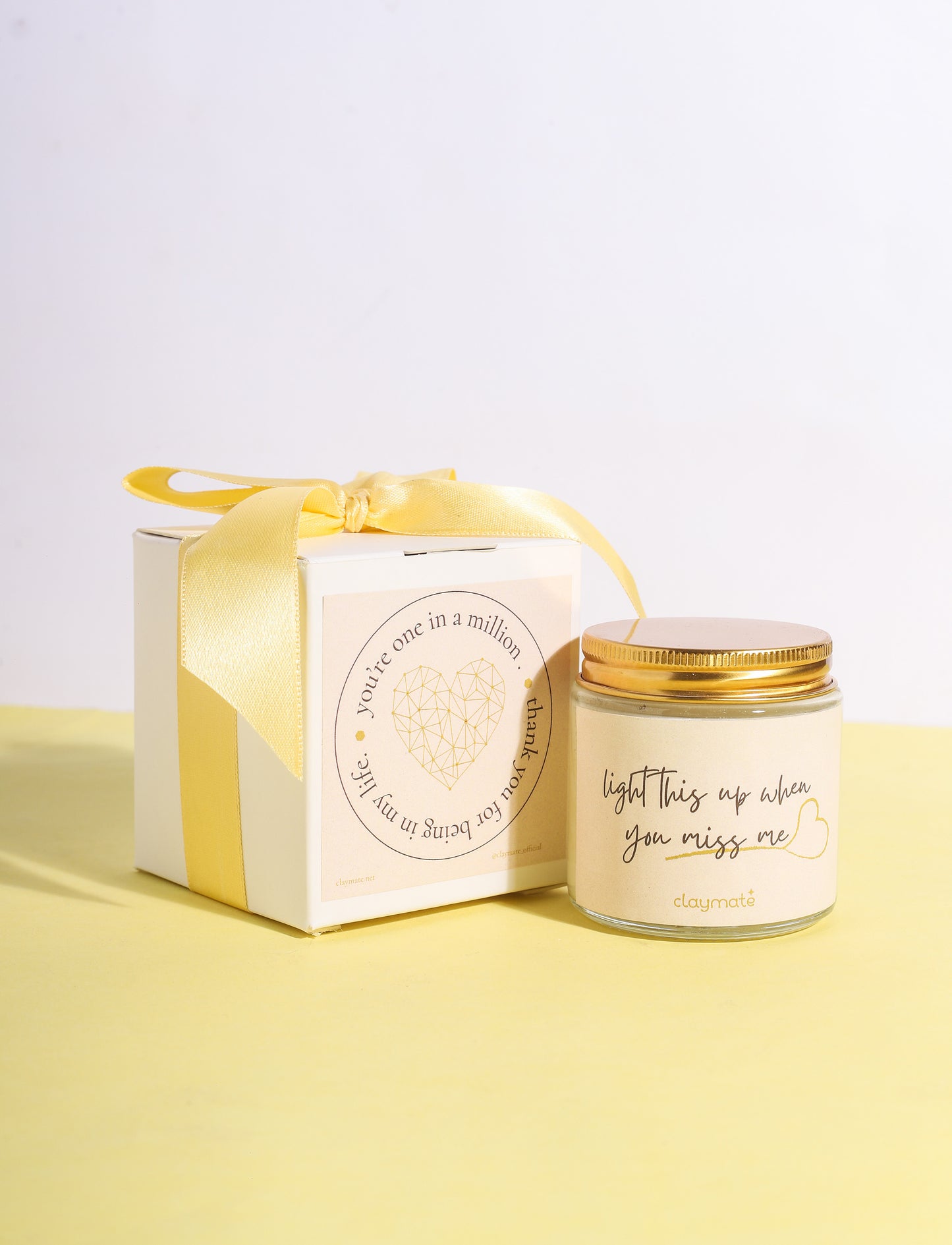 A candle with a beige label and a gold lid, displayed next to its packaging box which is also in beige with a yellow ribbon. The candle packaging has a printed message that reads 'Miss me?'.