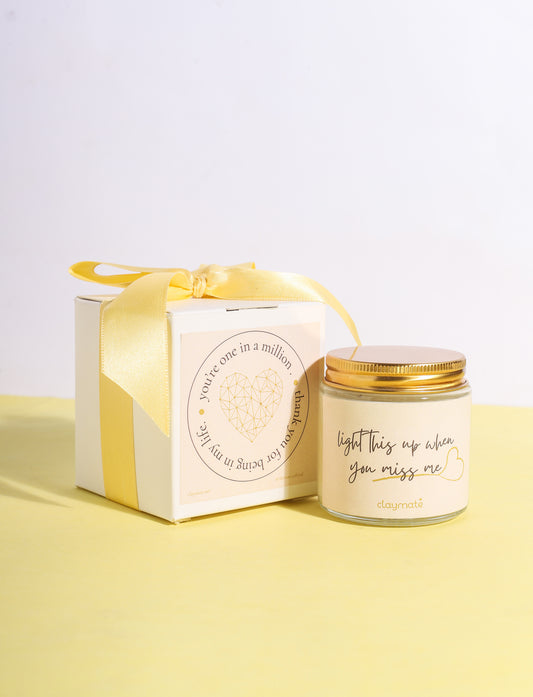 A candle with a beige label and a gold lid, displayed next to its packaging box which is also in beige with a yellow ribbon. The candle packaging has a printed message that reads 'Miss me?'.
