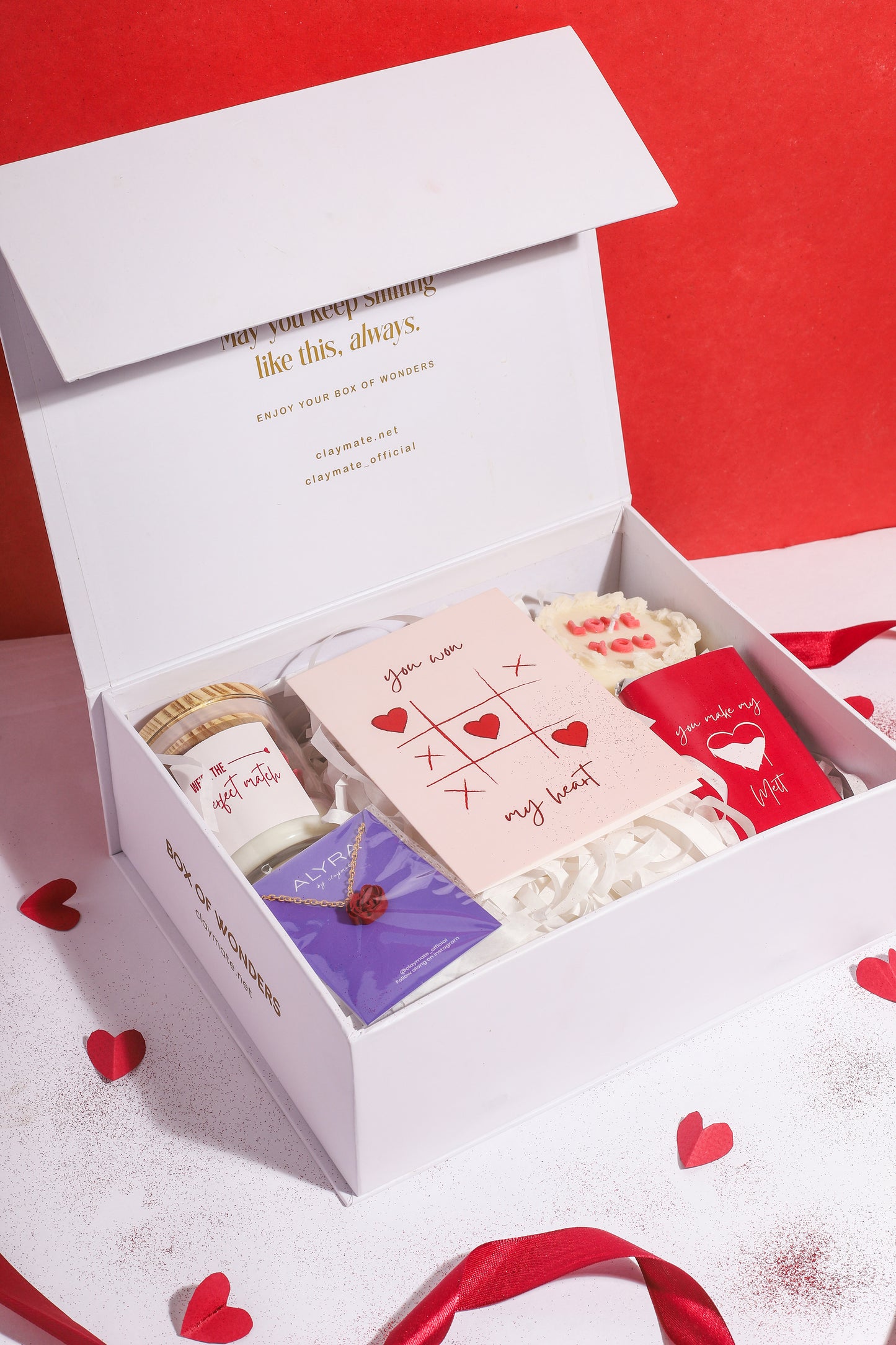 A gift box with a red ribbon, containing various items including a rosevel pendant, a wrapped chocolate bar, a red rose scented candle, a 'Love You' scented cake candle, and a cute card, all displayed on a white surface with red and white decorative hearts.