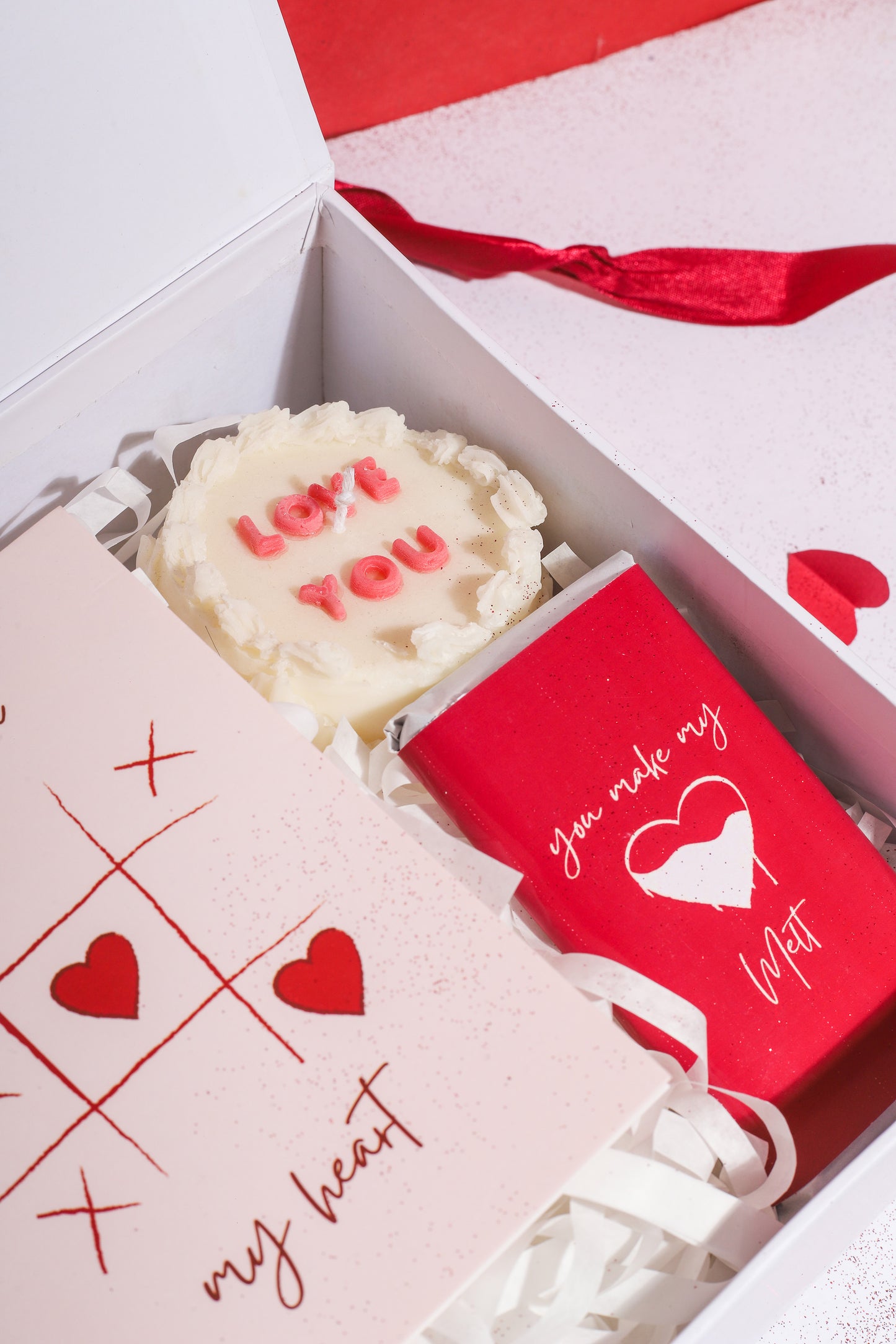 A gift box with a red ribbon, containing various items including a rosevel pendant, a wrapped chocolate bar, a red rose scented candle, a 'Love You' scented cake candle, and a cute card, all displayed on a white surface with red and white decorative hearts.