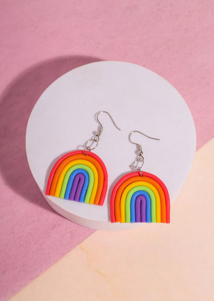 A pair of rainbow-shaped polymer clay earrings placed on a pink and beige background. The earrings feature layered arcs in red, orange, yellow, green, blue, and purple with silver hooks.