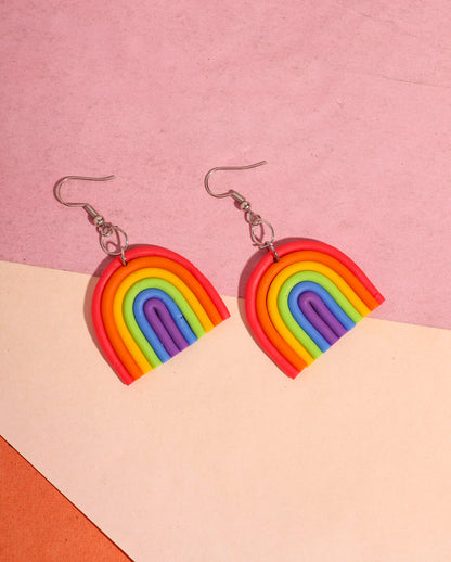 A pair of rainbow-shaped polymer clay earrings placed on a pink and beige background. The earrings feature layered arcs in red, orange, yellow, green, blue, and purple with silver hooks.