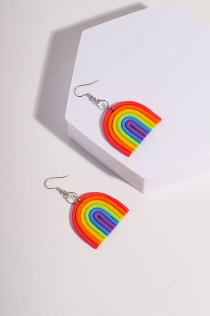 A pair of rainbow-shaped polymer clay earrings placed on a pink and beige background. The earrings feature layered arcs in red, orange, yellow, green, blue, and purple with silver hooks.