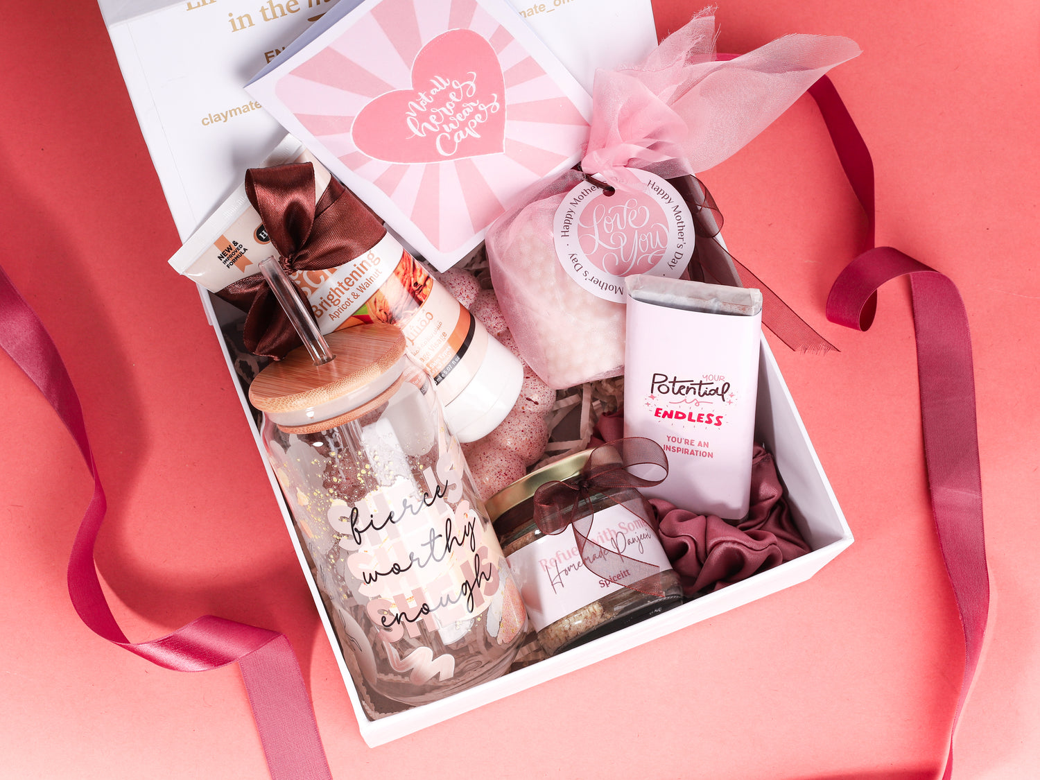 Empowerment-themed gift box featuring a pink chocolate bar with inspirational message, bamboo-lid glass tumbler with ‘fierce, worthy, enough’ quote, facial scrub tube, white bubble candle, jar of nuts, greeting card, and pink satin scrunchie on a peach background.