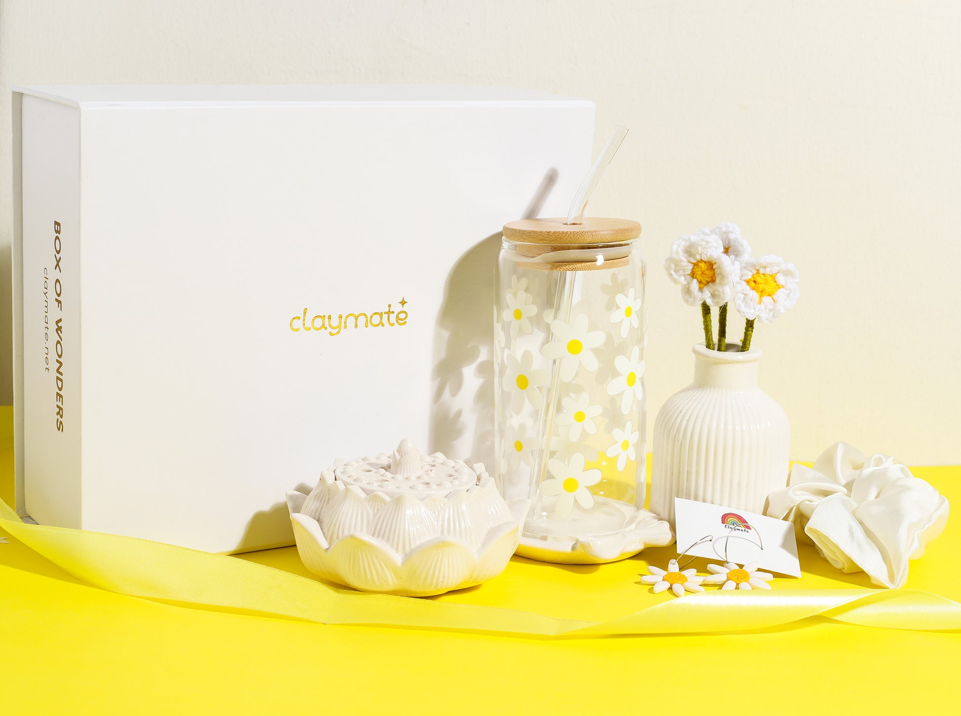 White gift box with yellow daisy-themed accessories including a glass tumbler with bamboo lid, crochet flower stems in a vase, ceramic trinket dish, daisy earrings, and a white scrunchie on a bright yellow background, Claymate gift set.
