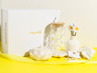 White gift box with yellow daisy-themed accessories including a glass tumbler with bamboo lid, crochet flower stems in a vase, ceramic trinket dish, daisy earrings, and a white scrunchie on a bright yellow background, Claymate gift set.
