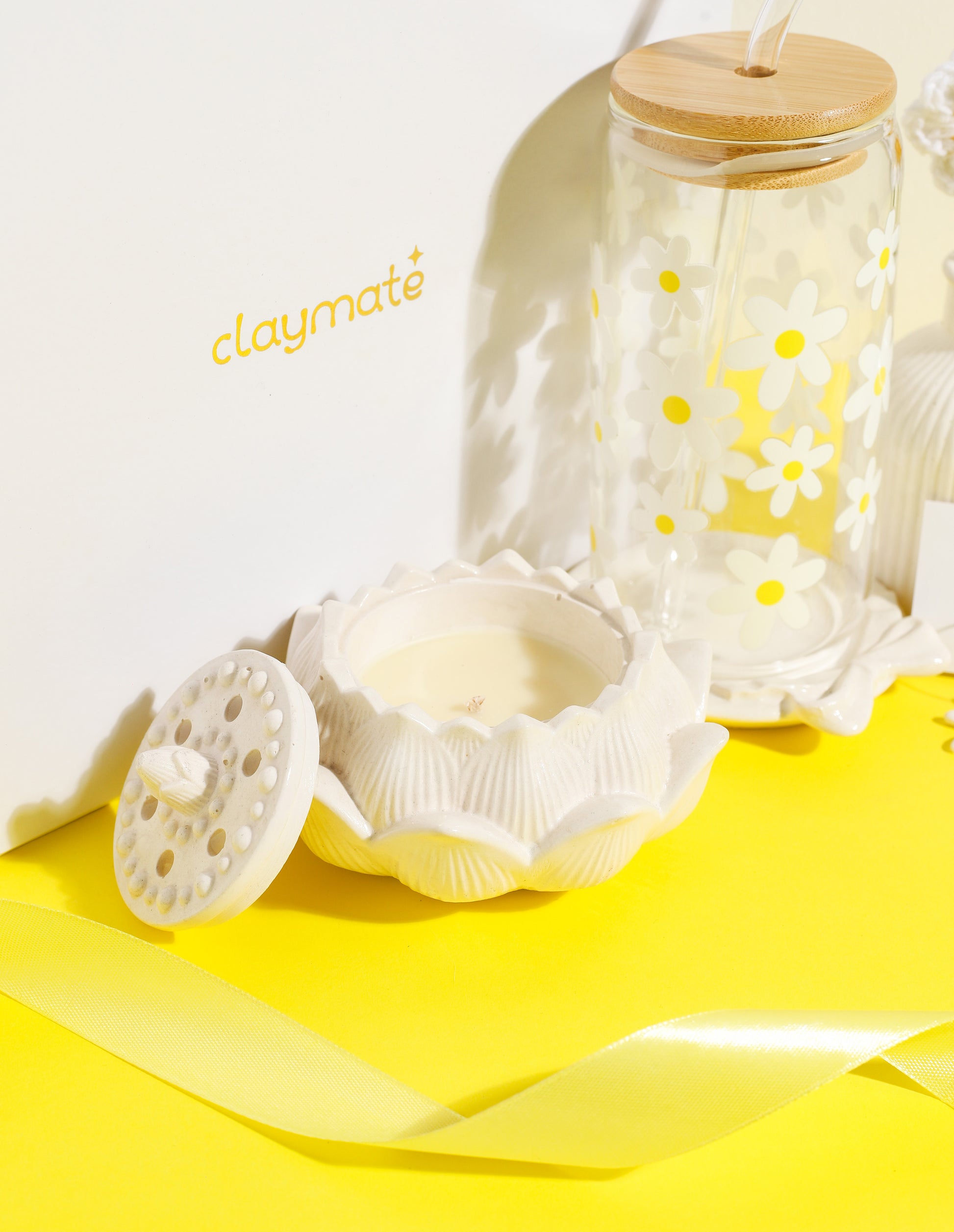 White gift box with yellow daisy-themed accessories including a glass tumbler with bamboo lid, crochet flower stems in a vase, ceramic trinket dish, daisy earrings, and a white scrunchie on a bright yellow background, Claymate gift set.