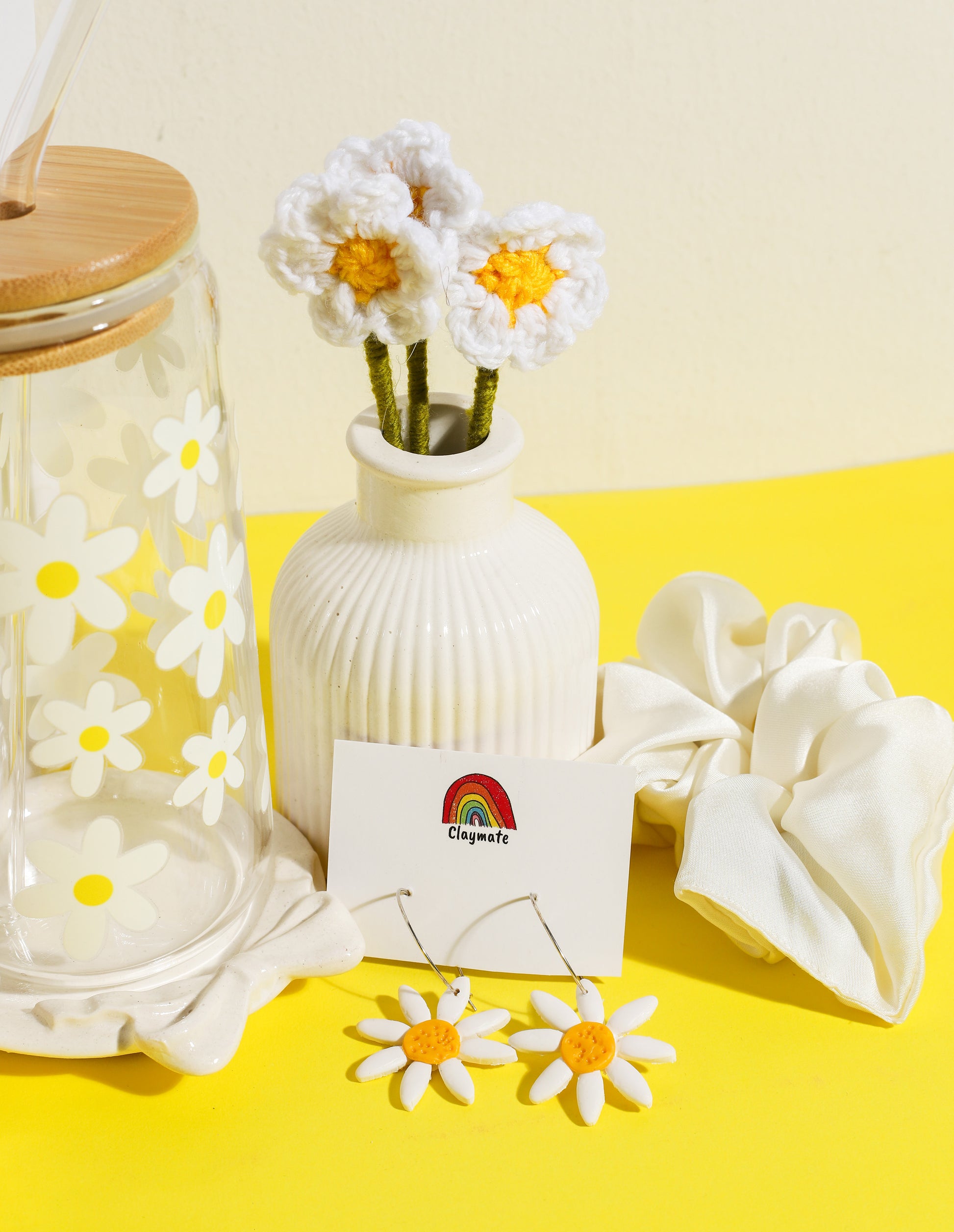 White gift box with yellow daisy-themed accessories including a glass tumbler with bamboo lid, crochet flower stems in a vase, ceramic trinket dish, daisy earrings, and a white scrunchie on a bright yellow background, Claymate gift set.