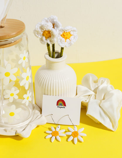 White gift box with yellow daisy-themed accessories including a glass tumbler with bamboo lid, crochet flower stems in a vase, ceramic trinket dish, daisy earrings, and a white scrunchie on a bright yellow background, Claymate gift set.