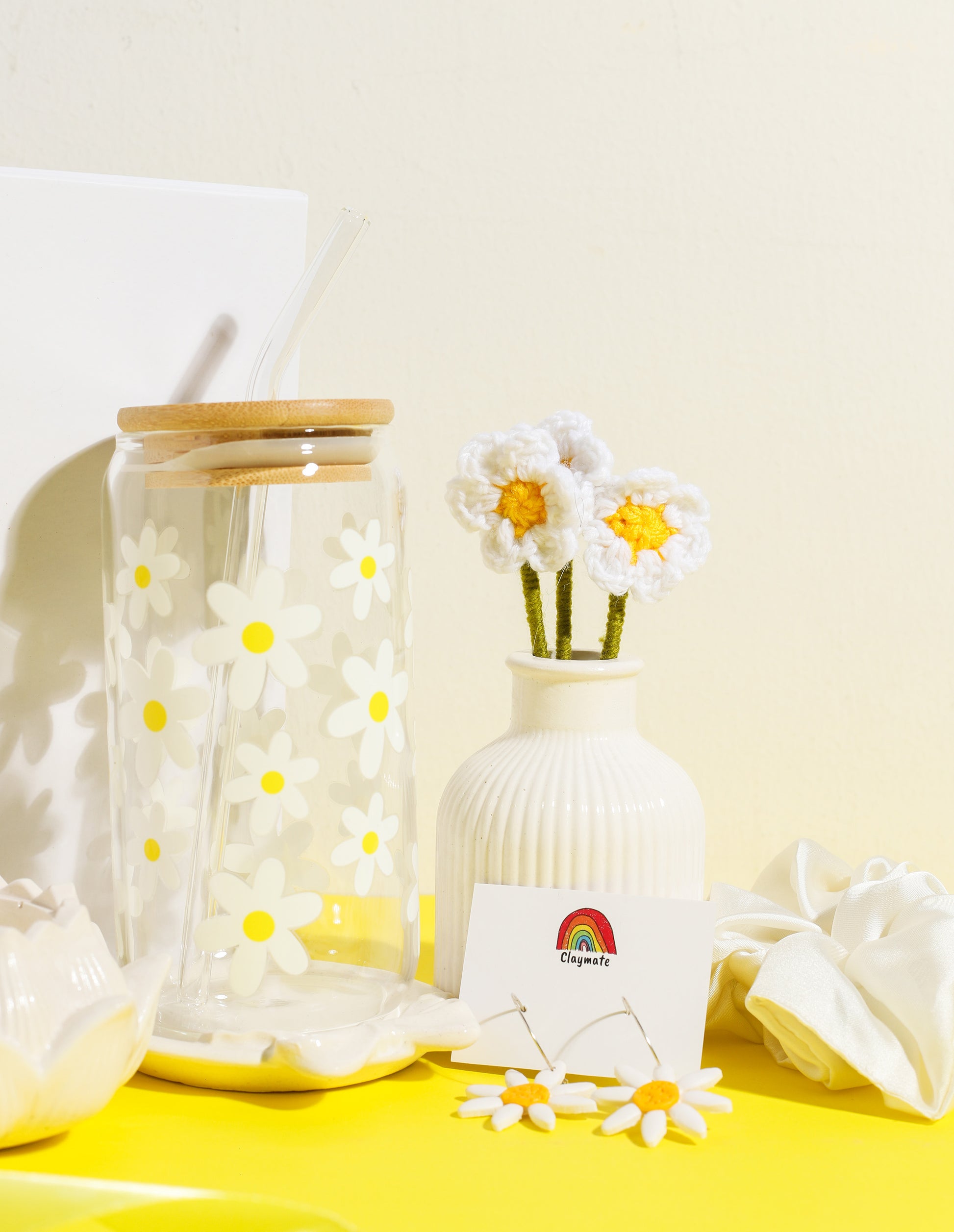 White gift box with yellow daisy-themed accessories including a glass tumbler with bamboo lid, crochet flower stems in a vase, ceramic trinket dish, daisy earrings, and a white scrunchie on a bright yellow background, Claymate gift set.