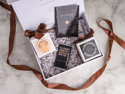 A gift box open to reveal its contents, which include a wrapped chocolate bar, a glass jar scented candle, a scent teddy bear candle, and a cute card, all arranged with a ribbon.