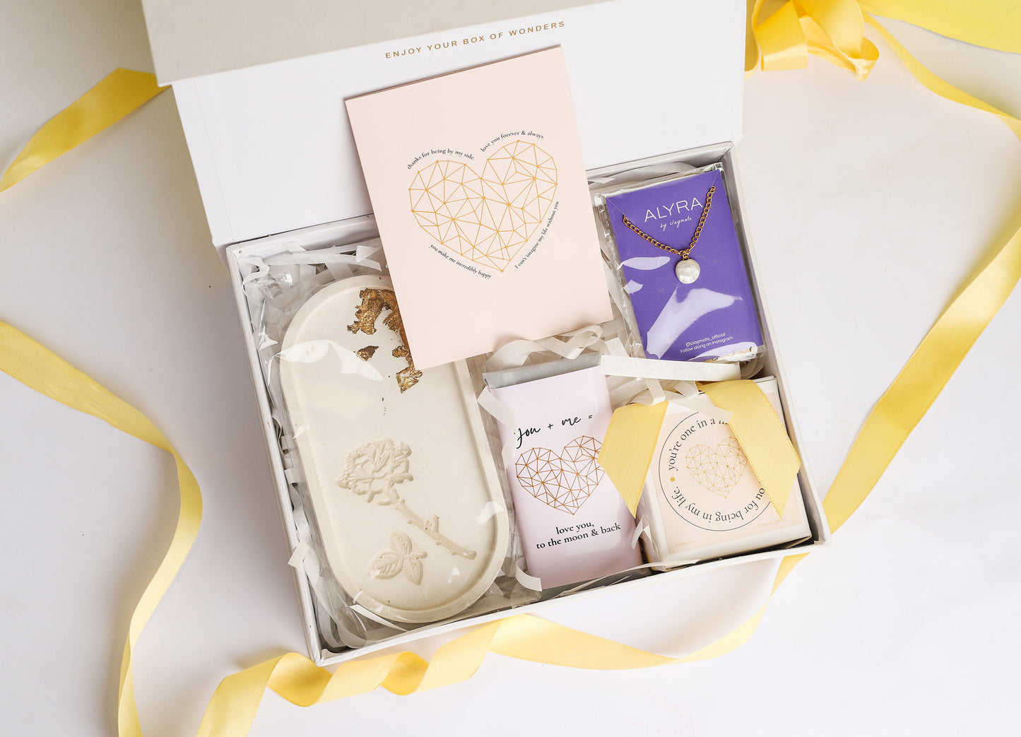 A gift box open to reveal its contents, which include a rose trinket tray, a pearl pendant in a black box, a wrapped chocolate bar, a glass jar scented candle, and a card. The box is adorned with a ribbon.