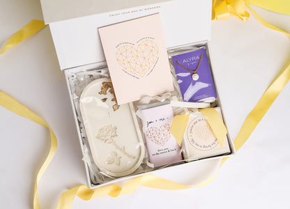 A gift box open to reveal its contents, which include a rose trinket tray, a pearl pendant in a black box, a wrapped chocolate bar, a glass jar scented candle, and a card. The box is adorned with a ribbon.