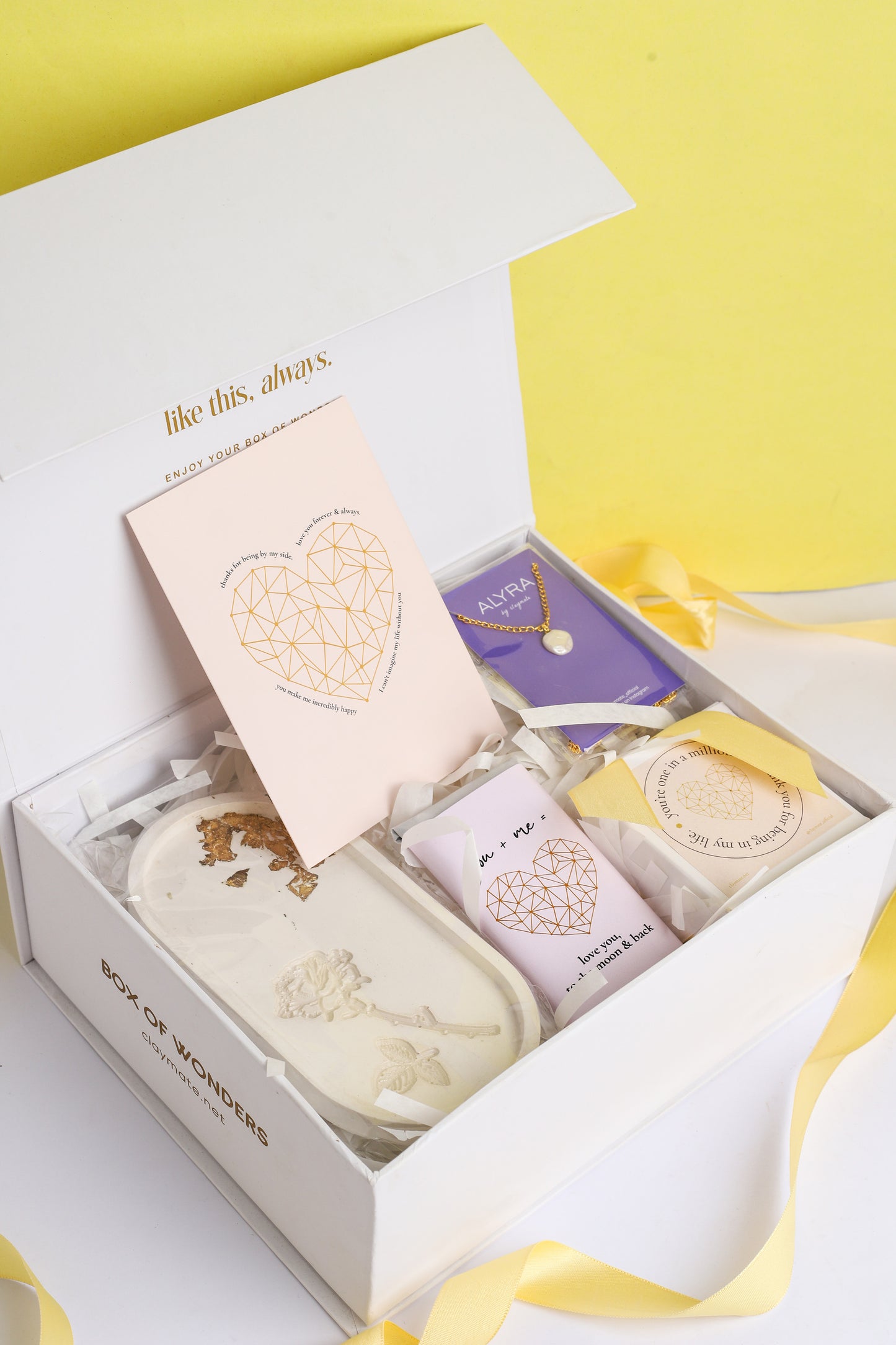 A gift box open to reveal its contents, which include a rose trinket tray, a pearl pendant in a black box, a wrapped chocolate bar, a glass jar scented candle, and a card. The box is adorned with a ribbon.