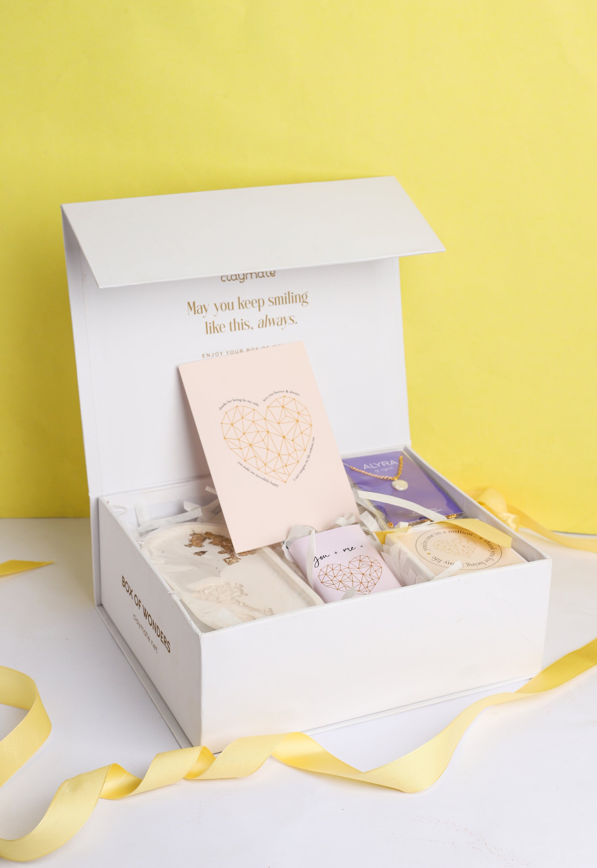 A gift box open to reveal its contents, which include a rose trinket tray, a pearl pendant in a black box, a wrapped chocolate bar, a glass jar scented candle, and a card. The box is adorned with a ribbon.