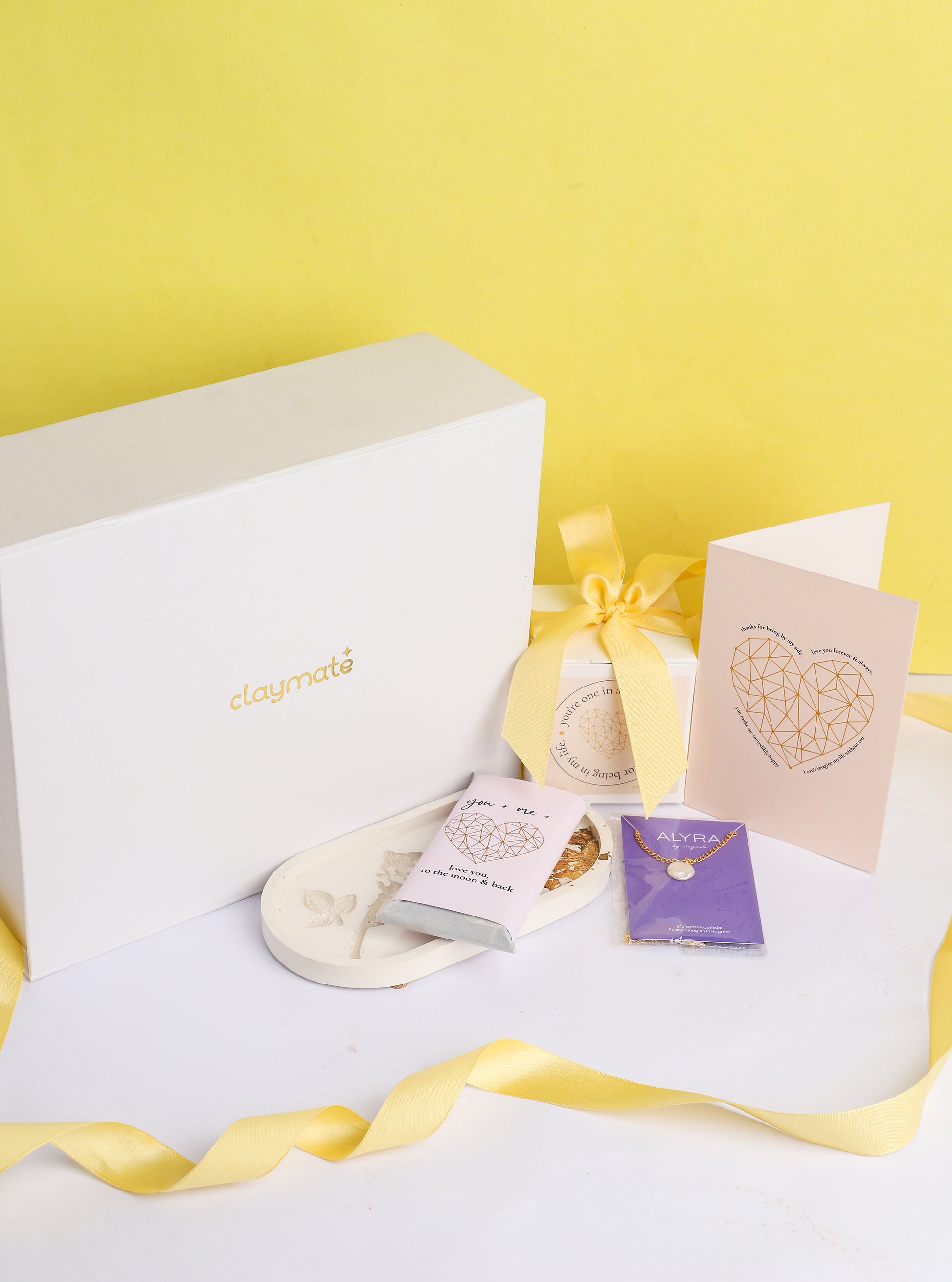 A gift box open to reveal its contents, which include a rose trinket tray, a pearl pendant in a black box, a wrapped chocolate bar, a glass jar scented candle, and a card. The box is adorned with a ribbon.