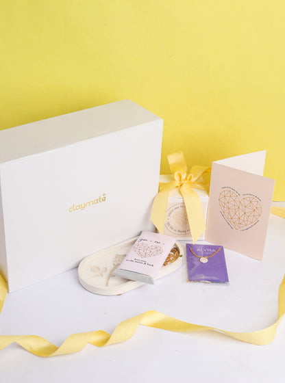 A gift box open to reveal its contents, which include a rose trinket tray, a pearl pendant in a black box, a wrapped chocolate bar, a glass jar scented candle, and a card. The box is adorned with a ribbon.