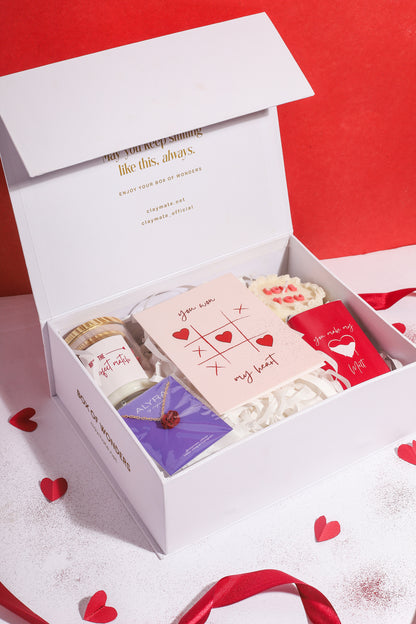 A gift box with a red ribbon, containing various items including a rosevel pendant, a wrapped chocolate bar, a red rose scented candle, a 'Love You' scented cake candle, and a cute card, all displayed on a white surface with red and white decorative hearts.