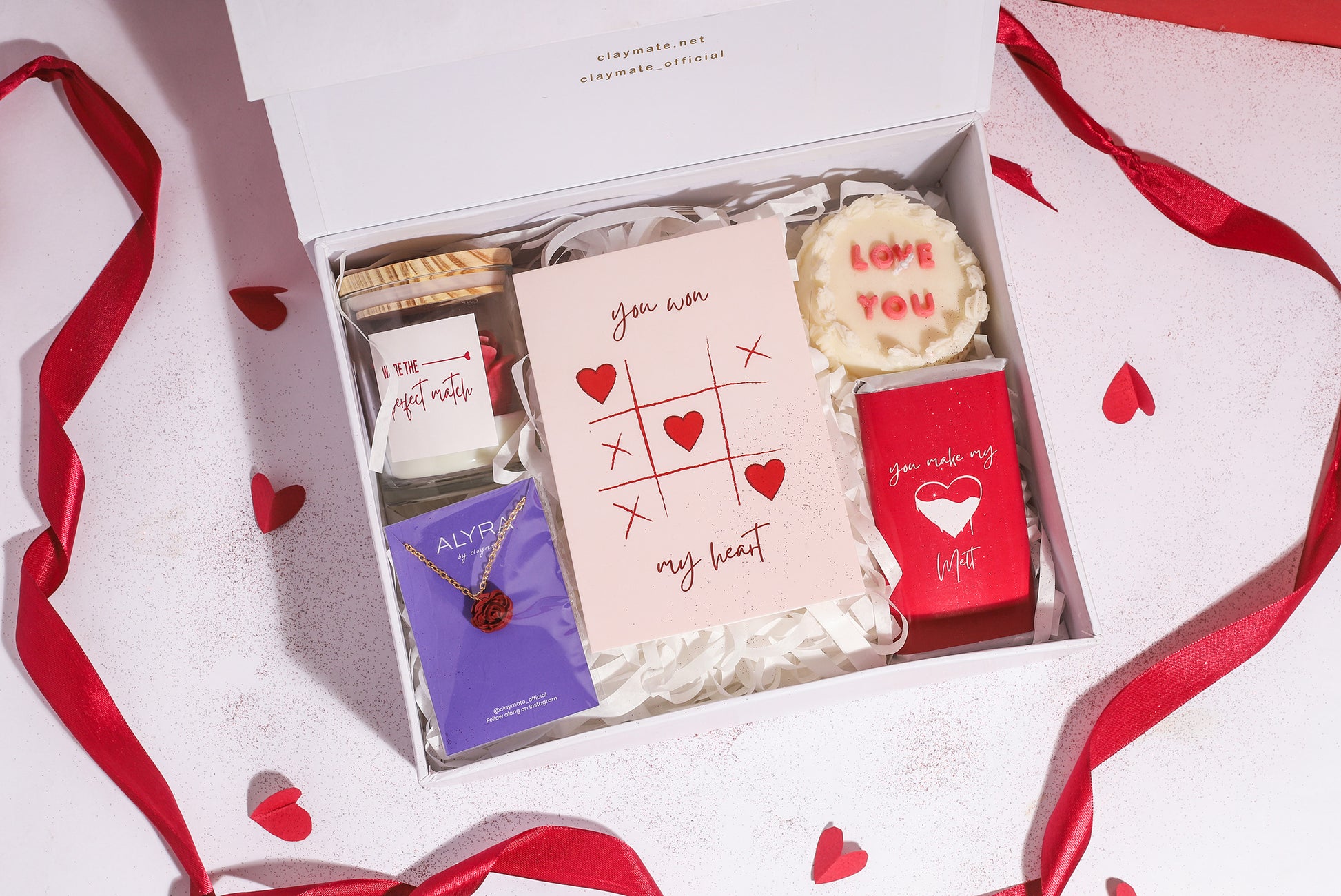 A gift box with a red ribbon, containing various items including a rosevel pendant, a wrapped chocolate bar, a red rose scented candle, a 'Love You' scented cake candle, and a cute card, all displayed on a white surface with red and white decorative hearts.
