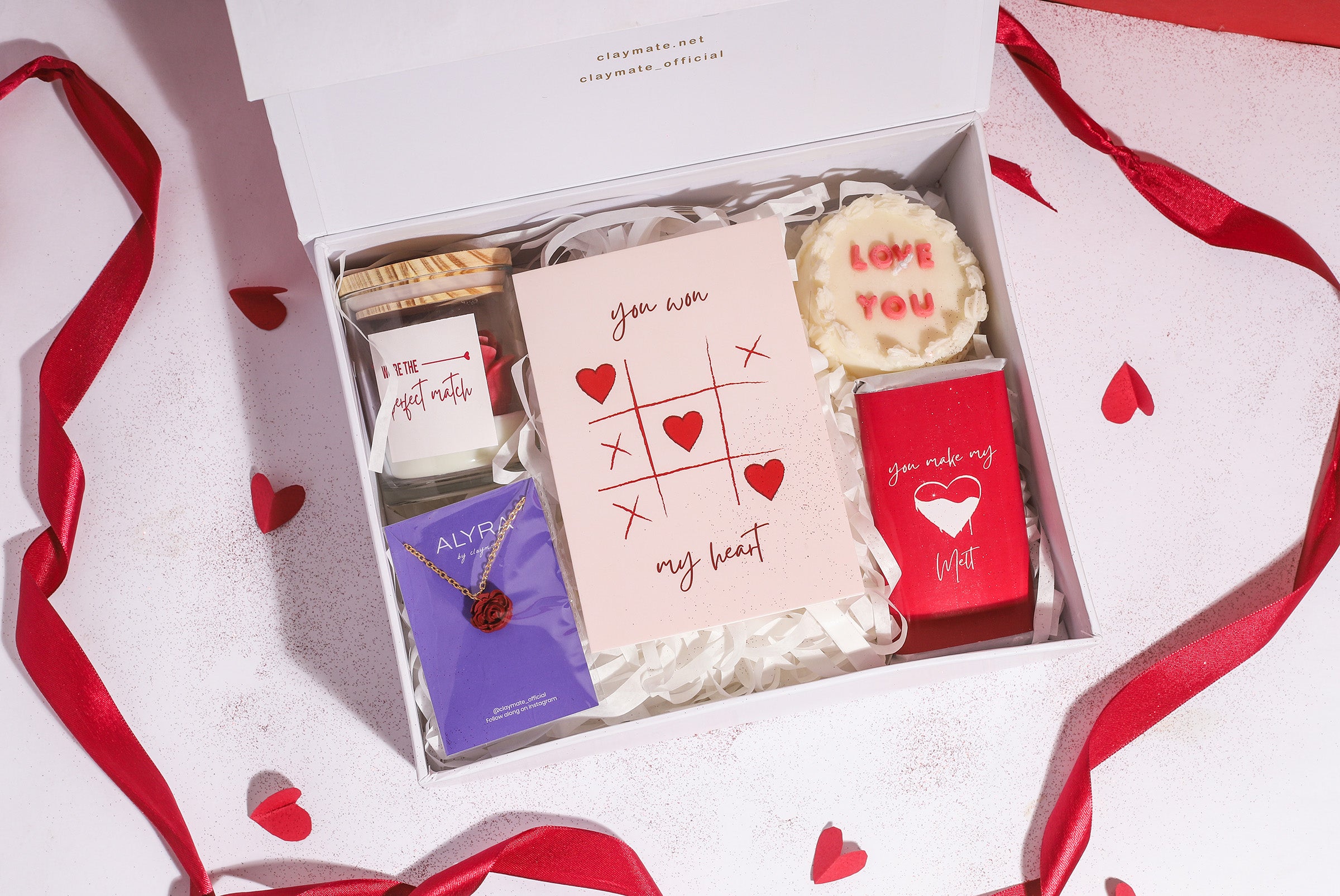 A gift box with a red ribbon, containing various items including a rosevel pendant, a wrapped chocolate bar, a red rose scented candle, a 'Love You' scented cake candle, and a cute card, all displayed on a white surface with red and white decorative hearts.
